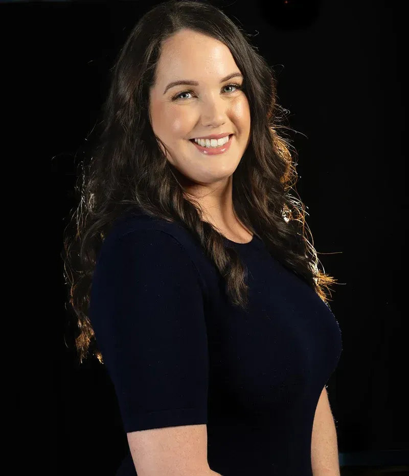 Smiling woman with long wavy brown hair in a dark navy dress against a black background