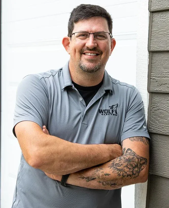 Smiling man in a gray polo shirt, arms crossed, standing beside a stone wall outdoors.