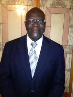 Man in a navy suit and striped tie standing against a tiled wall, smiling at the camera.