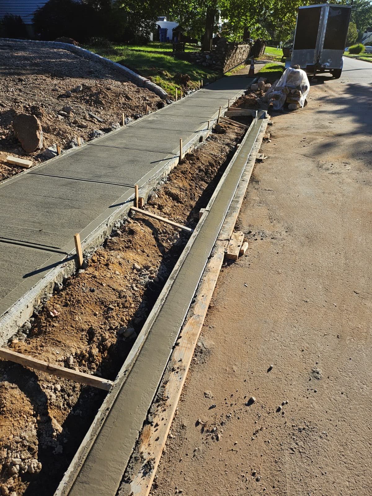 Sidewalk under construction beside a road, with fresh concrete curbs and dirt excavation.