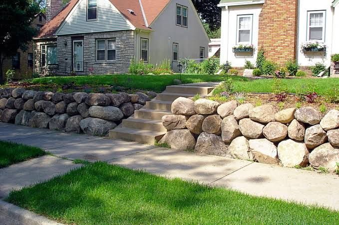 Stone retaining walls and steps beside a green lawn in front of suburban houses.