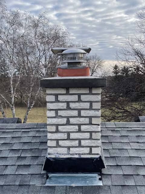 Brick chimney with metal cap on a gray shingled roof against cloudy sky and bare trees