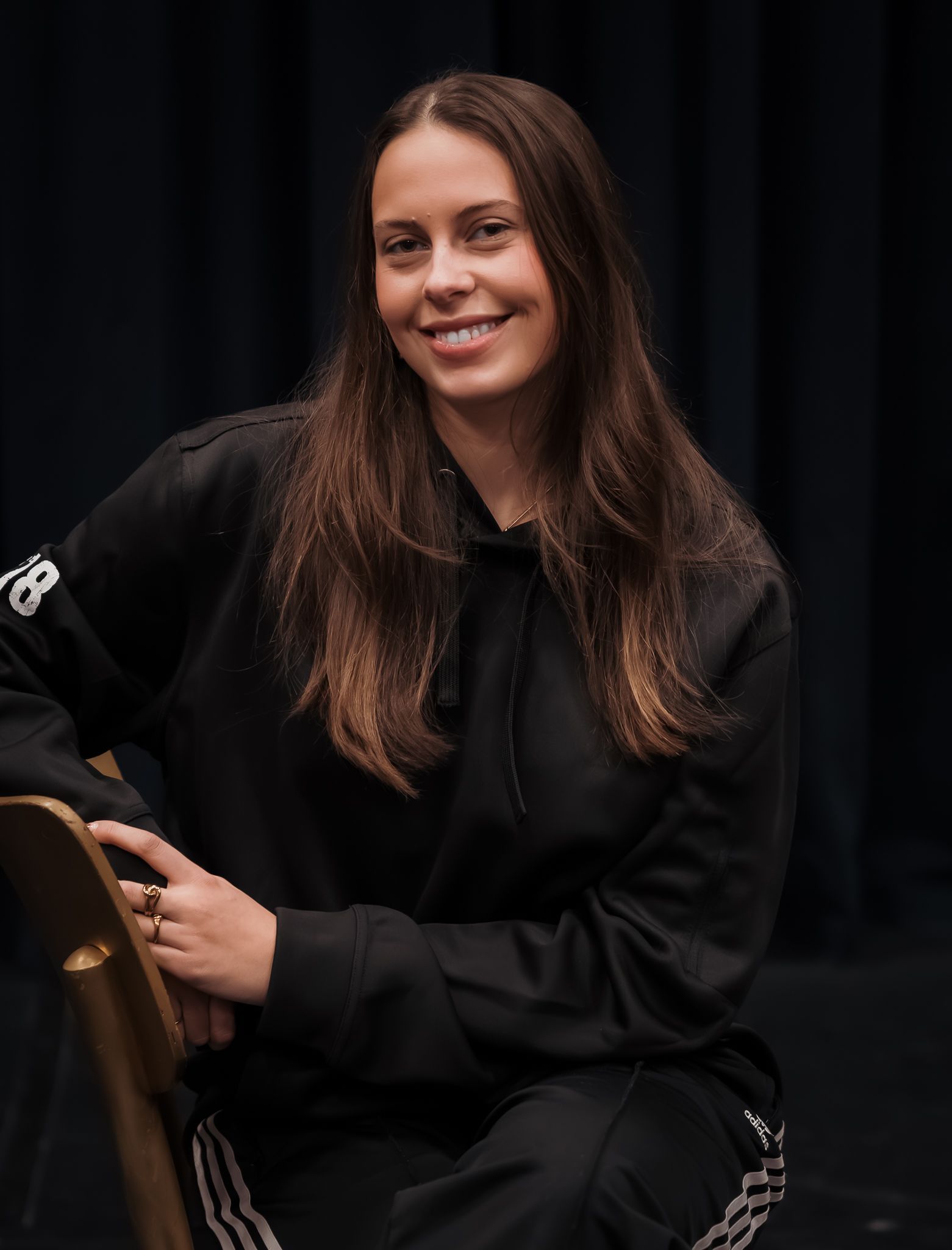 Een vrouw in een zwarte hoodie glimlacht, zittend op een stoel tegen een donkere achtergrond.
