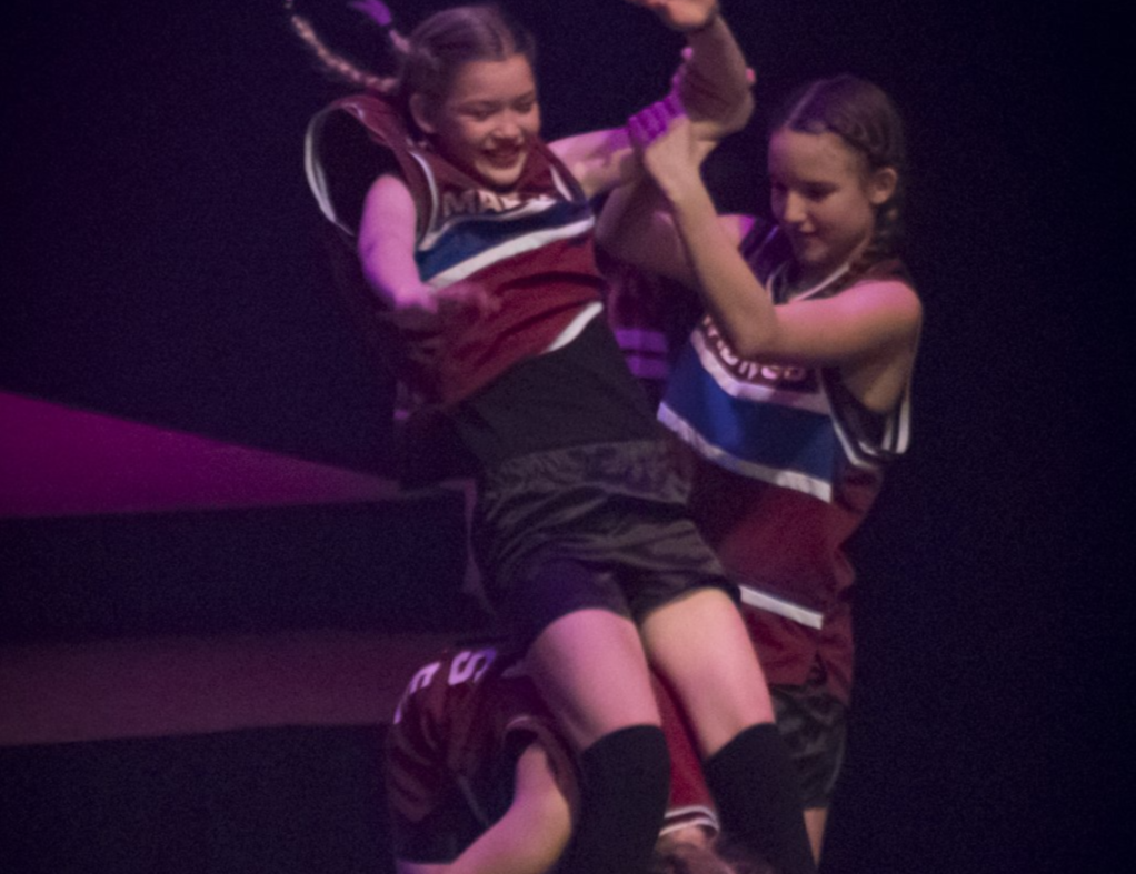 Cheerleaders voeren een stunt uit: een van hen wordt lachend opgetild door anderen in bordeauxrode en zwarte uniformen.