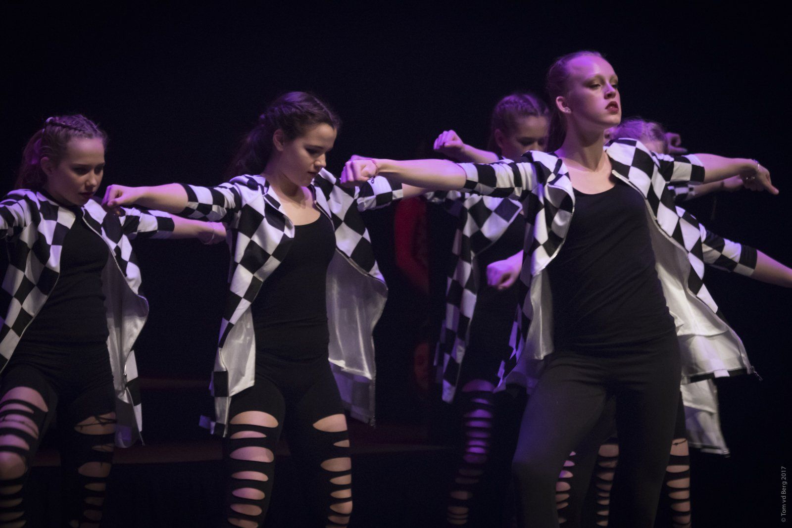 Dansers voeren een routine uit op het podium. Ze dragen zwarte outfits met geruite jasjes en staan ​​met uitgestrekte armen in een pose.