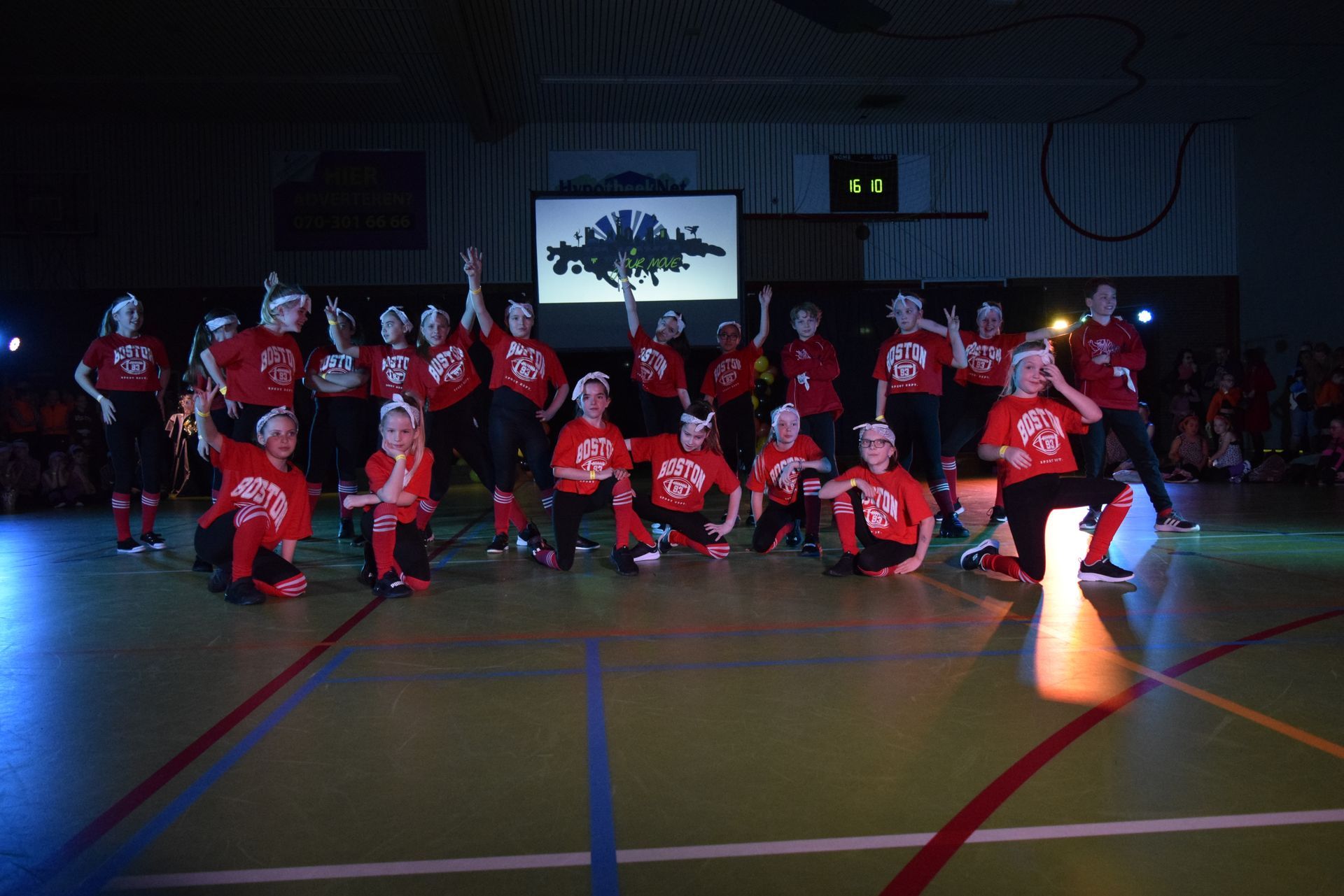 Een groep dansers in rode shirts en zwarte broeken treedt op in een gymzaal met podiumverlichting.