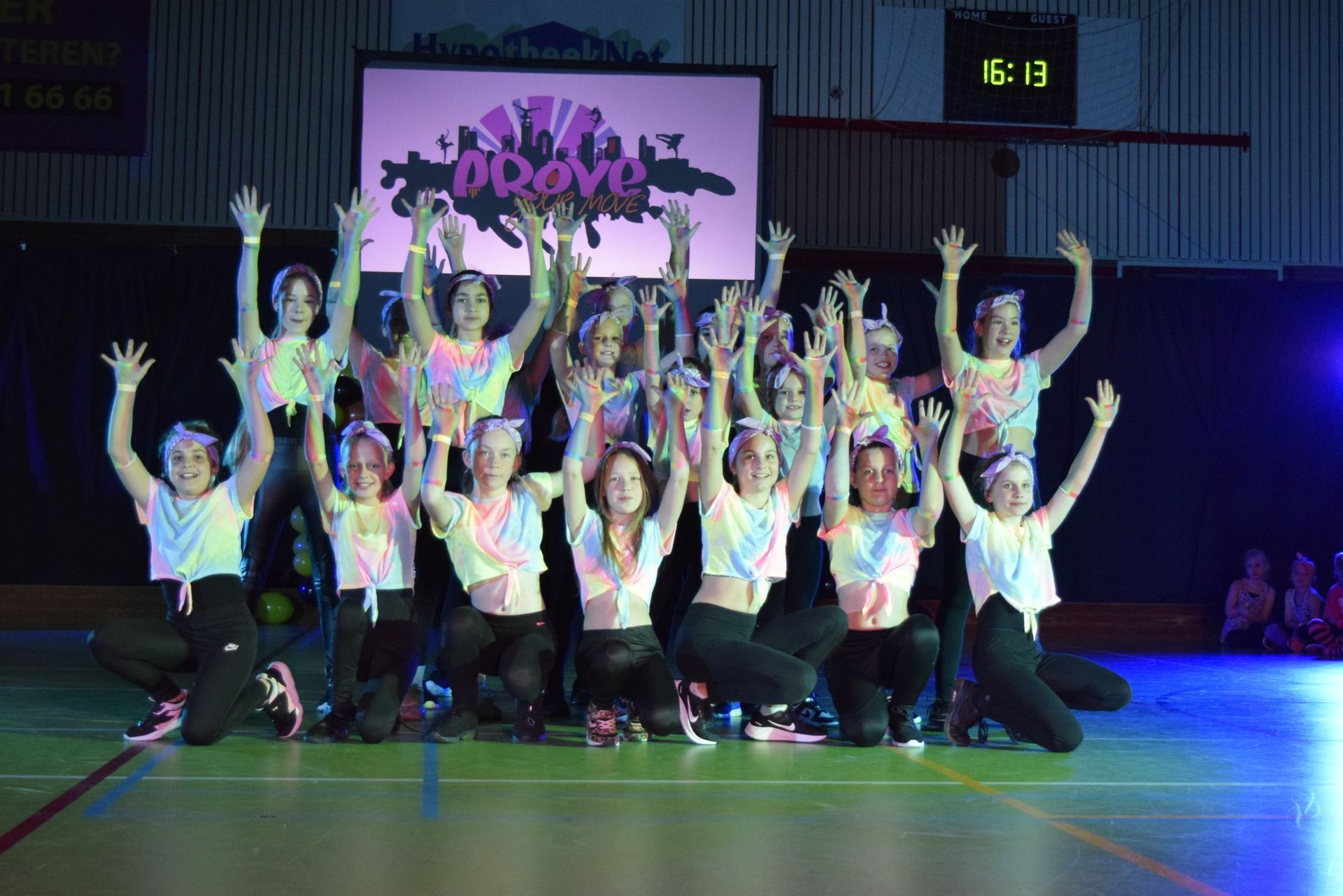Een groep dansers in kleurrijke tie-dye shirts en zwarte broeken, met de armen omhoog, treedt op een podium op.