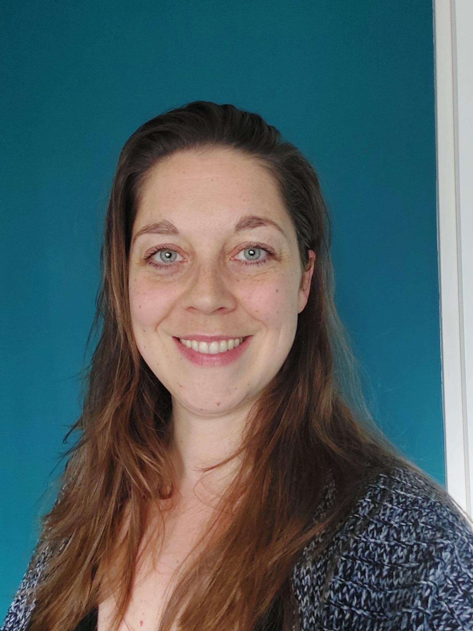 Lachende vrouw met lang bruin haar en blauwe ogen, tegen een turquoise muur op de achtergrond.