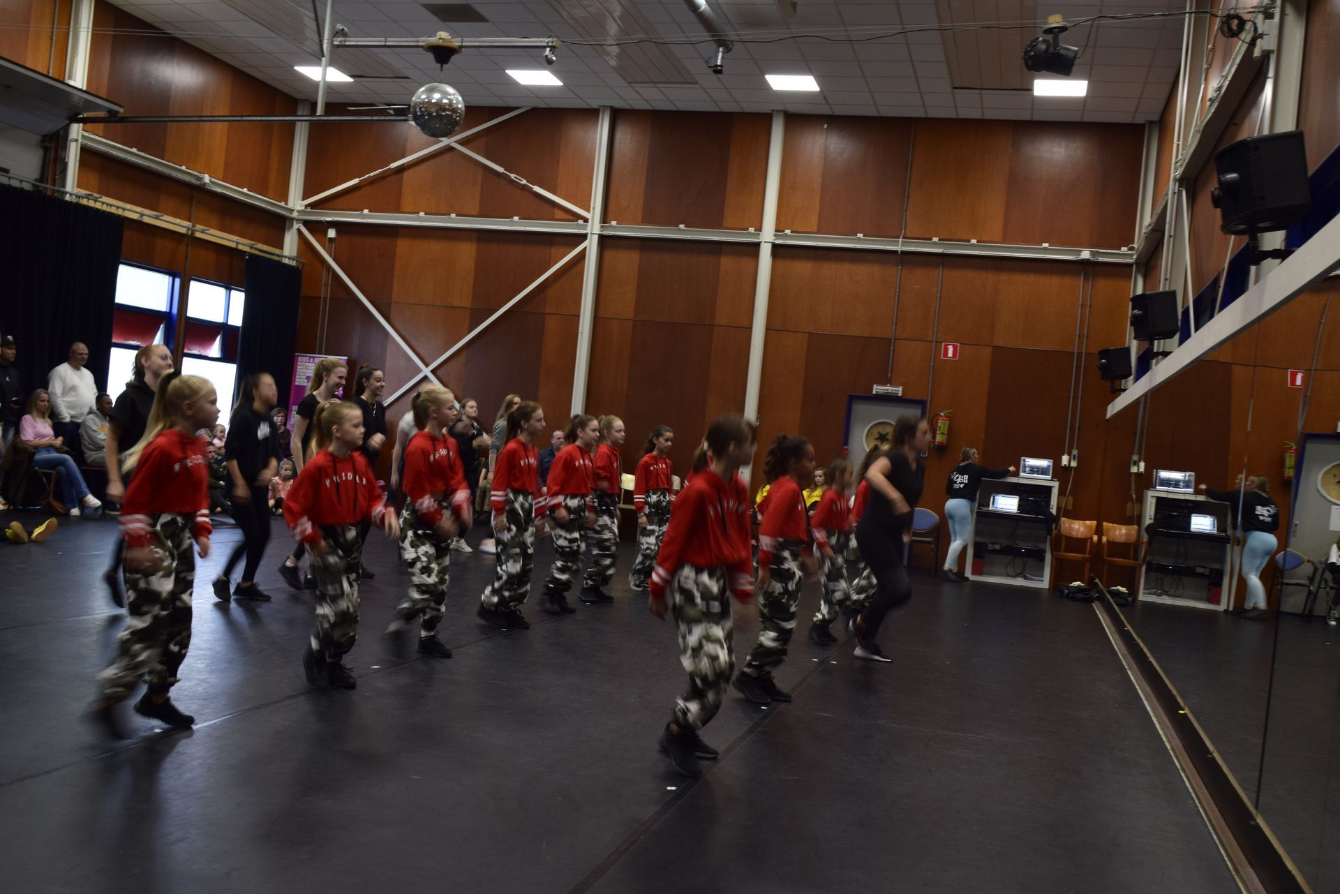Een groep kinderen in rode shirts en camouflagebroeken danst in een studio.