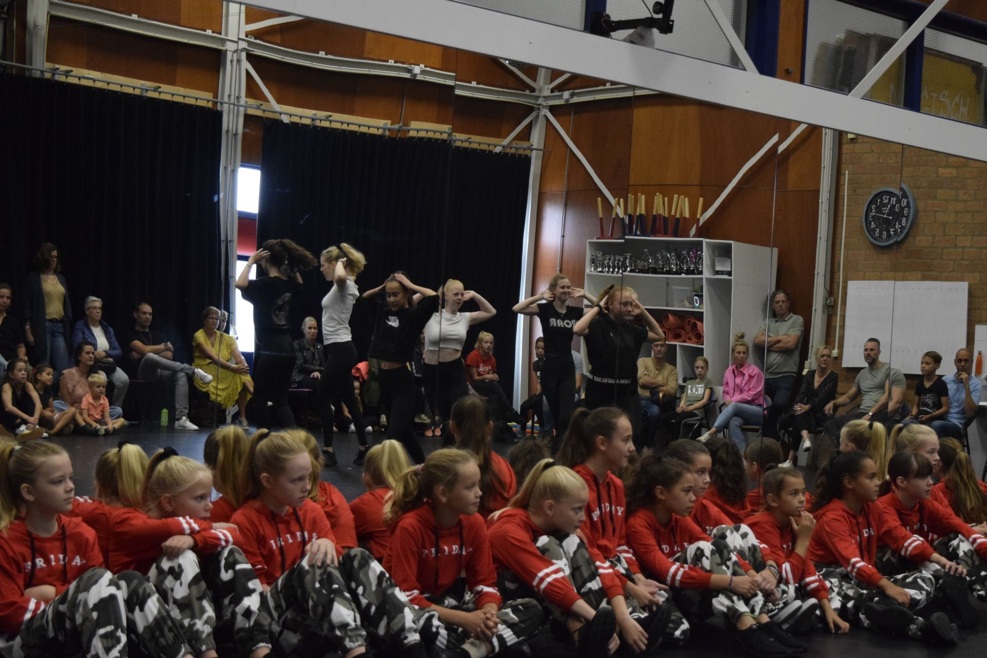 Dansers treden op het podium op, met publiek op de stoelen; sommigen dragen rood, anderen casual kleding.