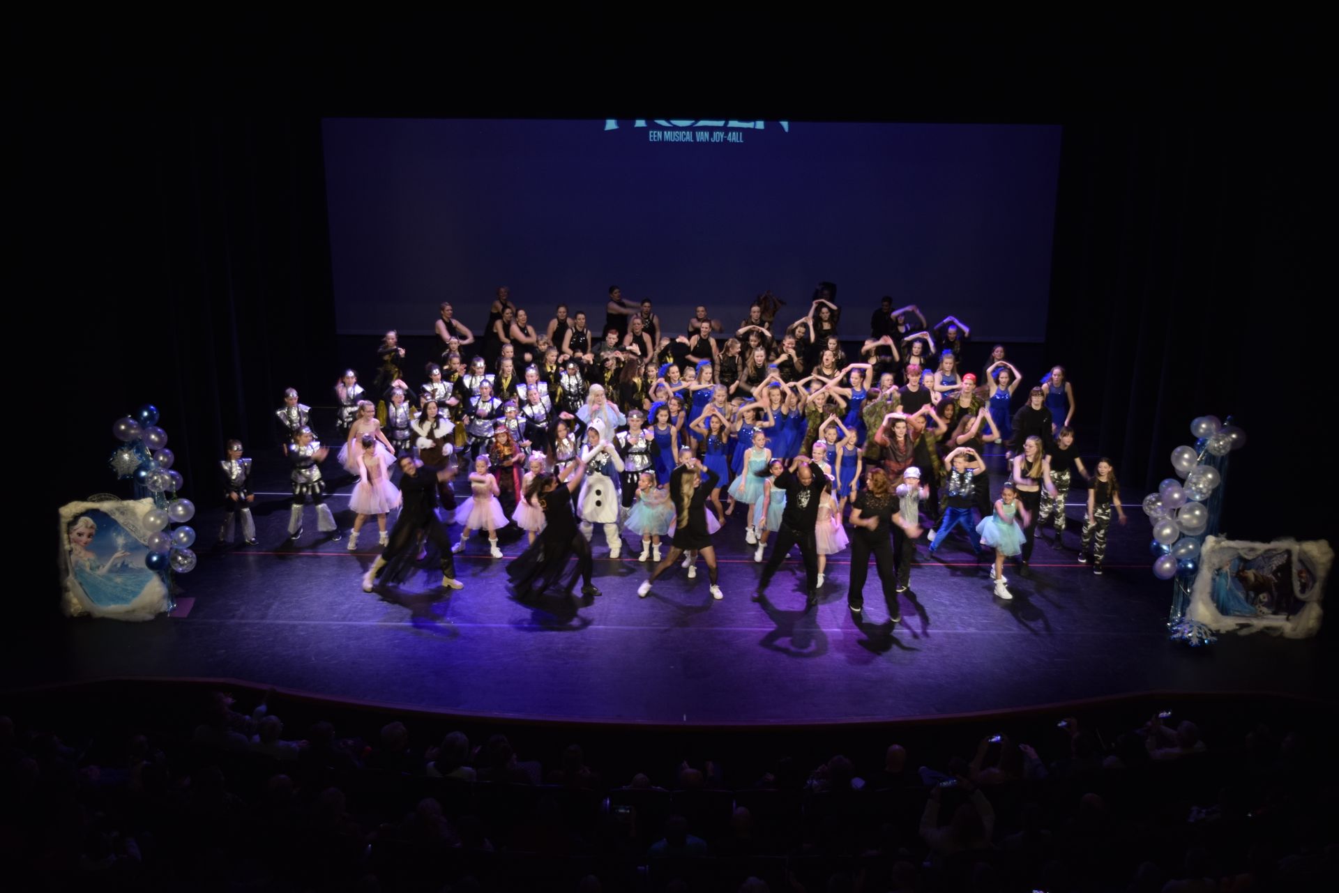 Dansers op het podium, velen in tutu's en kostuums, poseren onder een donkerblauw scherm. Ballonnen versieren de zijkant van het podium.