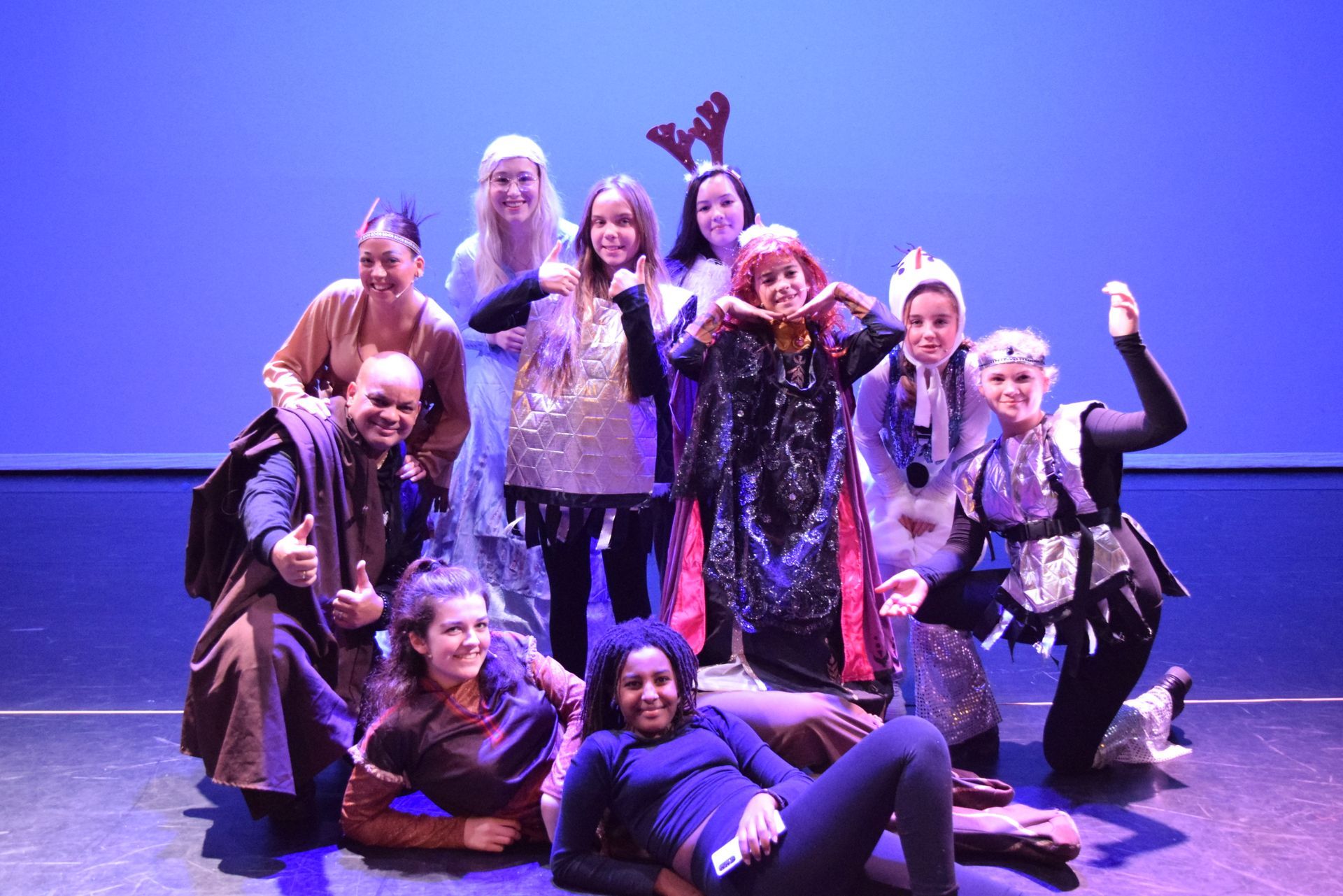 Een groep artiesten in kostuums poseert op een podium met een blauwe achtergrond. Ze glimlachen en maken speelse gebaren.