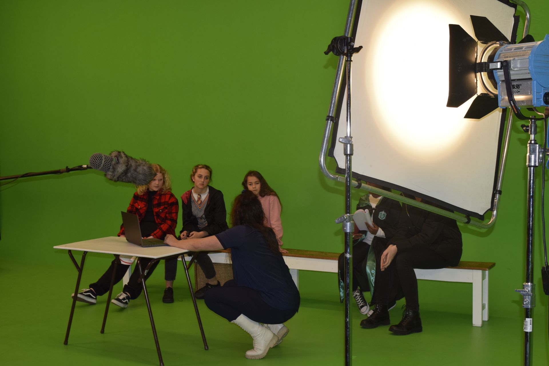 Mensen filmen voor een groen scherm. Een crewlid werkt aan een laptop aan een tafel. Lampen en een microfoon zijn zichtbaar.