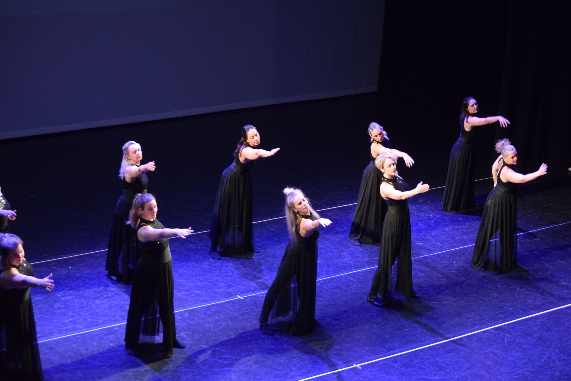 Dansers in zwarte jurken treden op het podium op, met uitgestrekte armen.