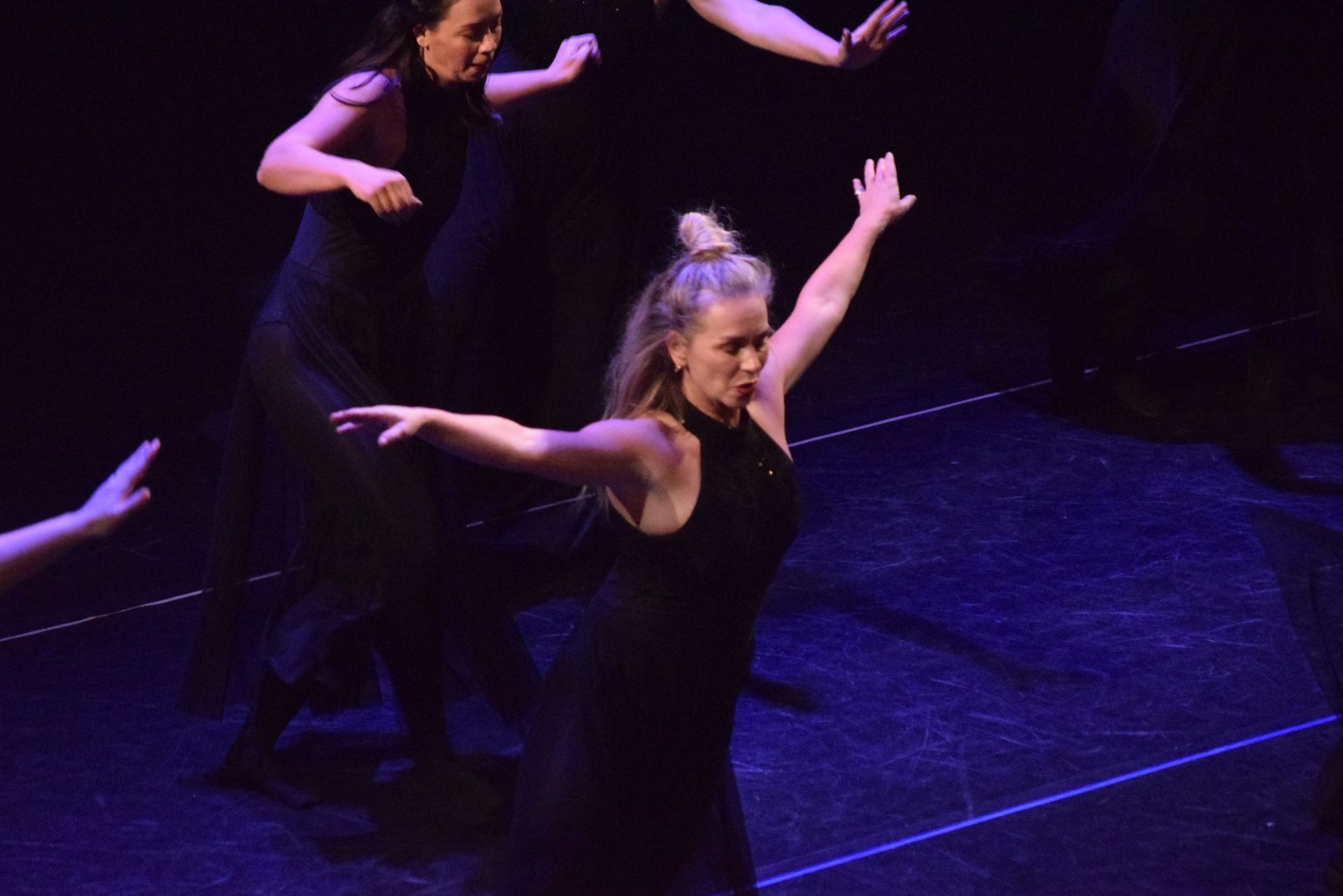 Danseressen in zwarte jurken treden op een donker podium op met blauw licht. Een van de danseressen heeft haar armen uitgestrekt.