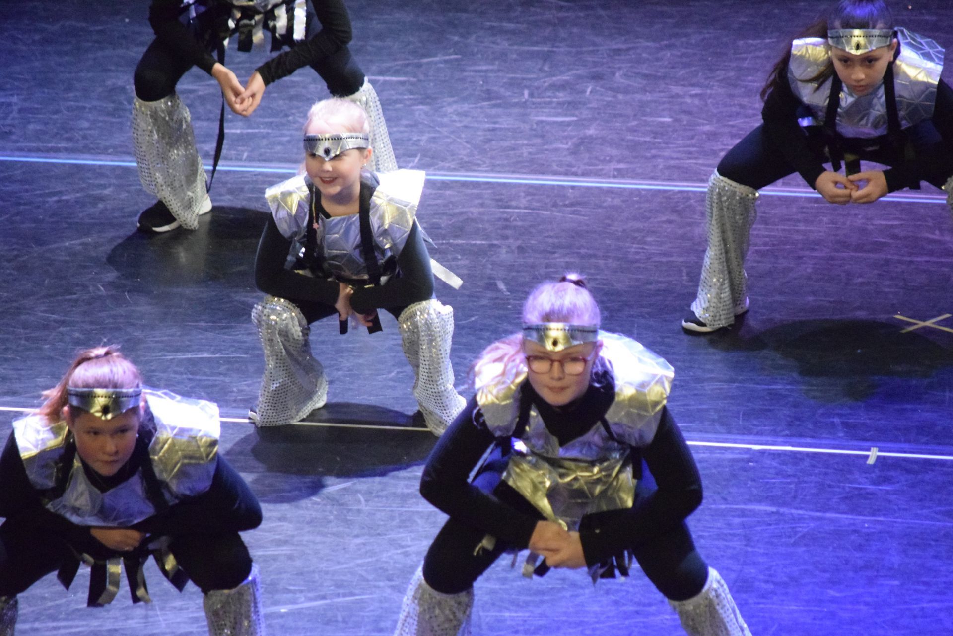 Dansers in futuristische kostuums treden op een podium op, vastberaden gehurkt. Zilver en zwart.