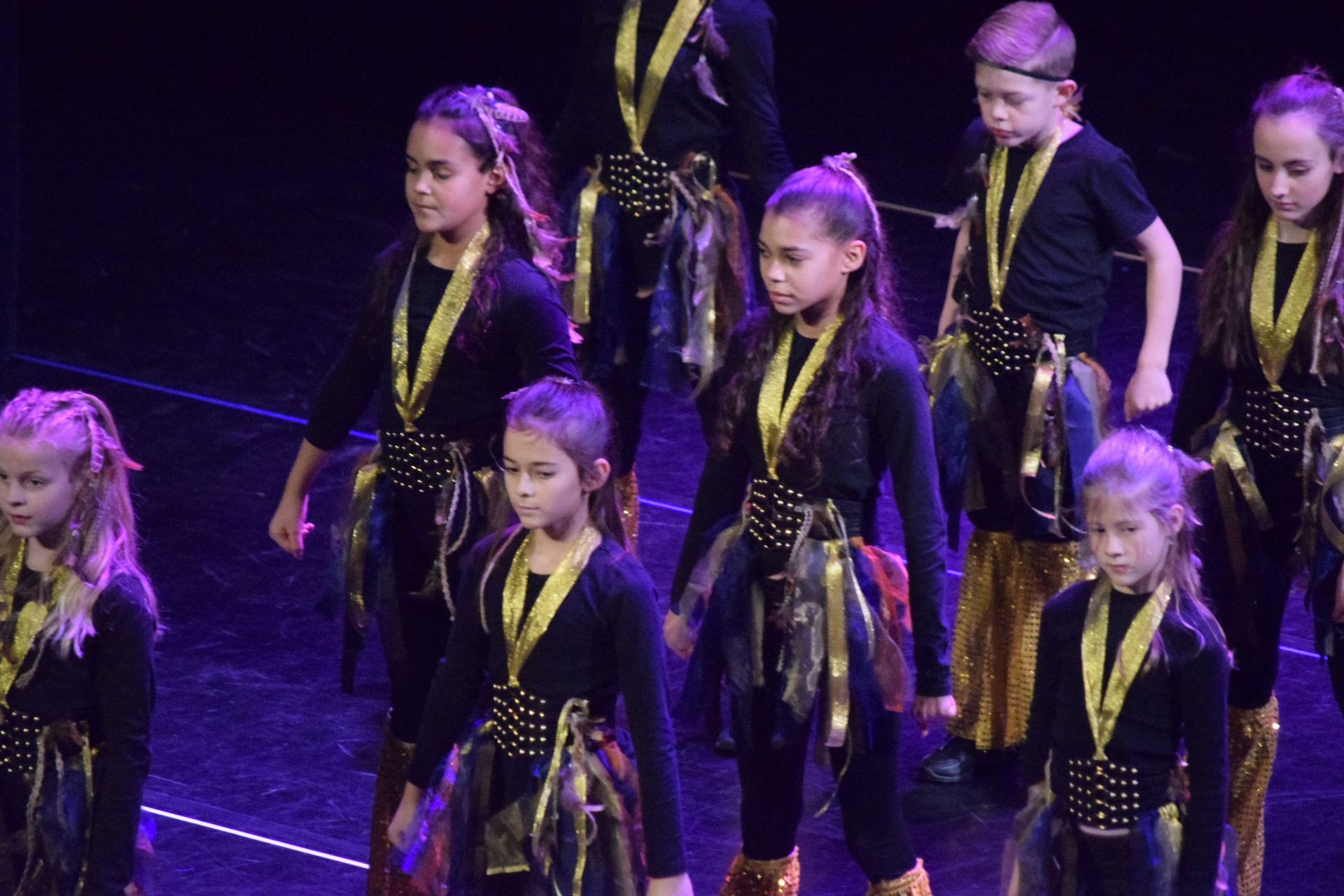 Kinderen in zwart-gouden kostuums op een podium. Ze staan ​​in een rij en kijken vooruit.