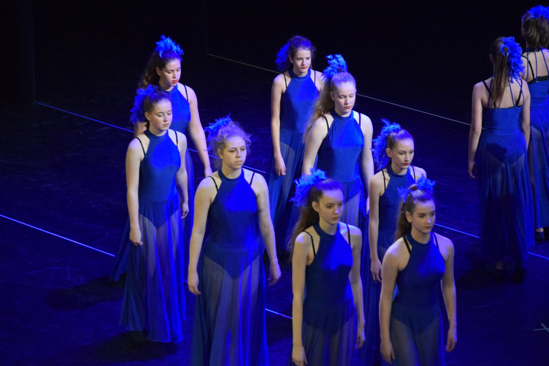 Danseressen in blauwe jurken op het podium, met donker haar, sommigen met verenaccessoires.