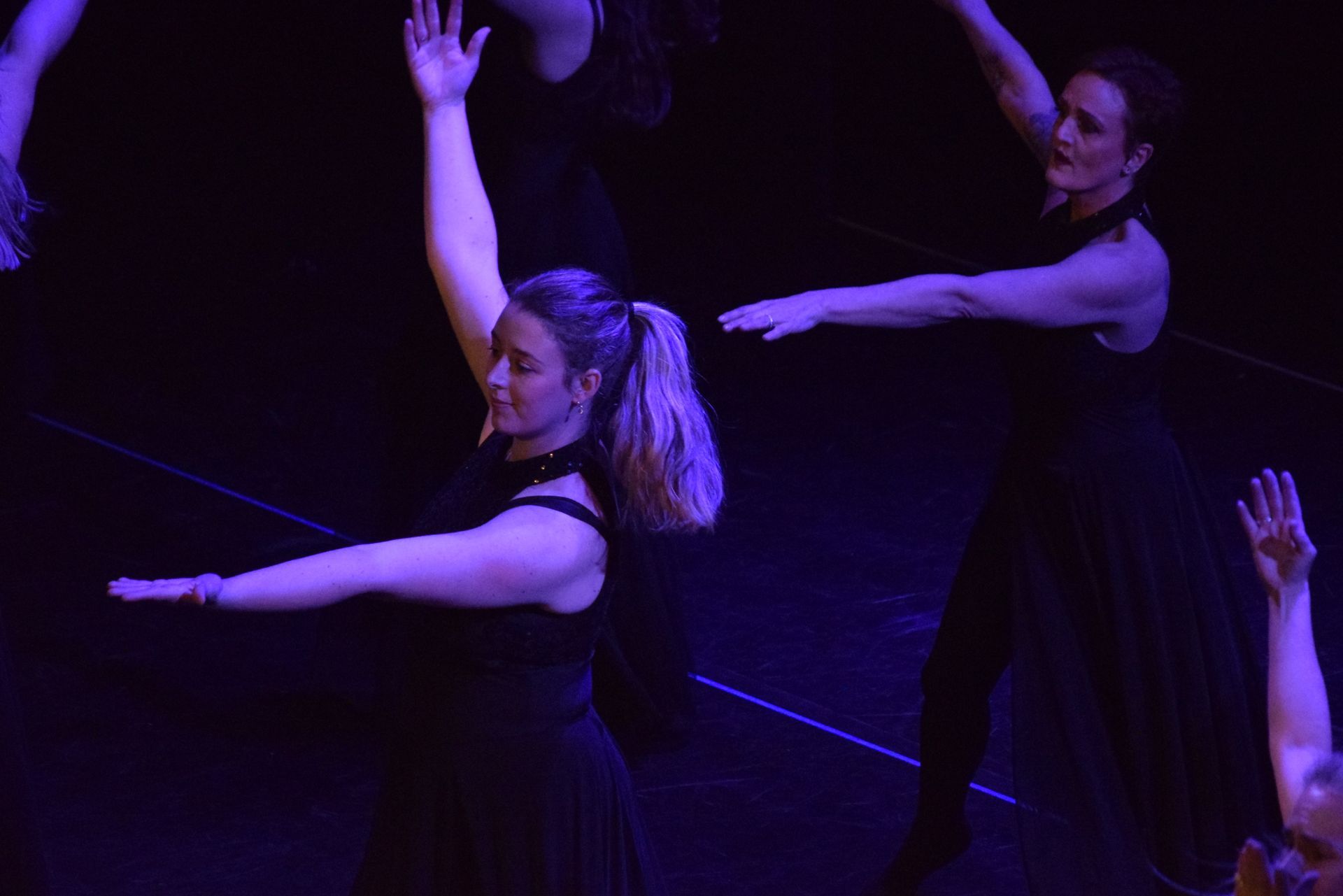 Dansers in donkere kleding op een podium met paarse verlichting, met uitgestrekte armen tijdens een optreden.