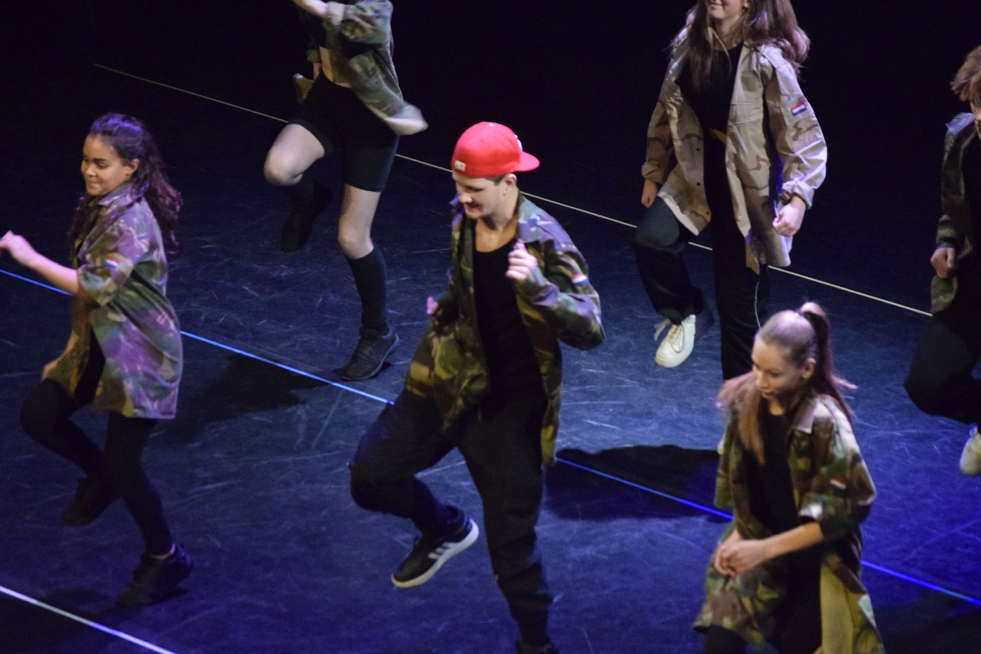 Dansers voeren een hiphoproutine uit op het podium. De meesten dragen camouflagejassen. Een man met een rode pet danst in het midden. Het podium is donker.