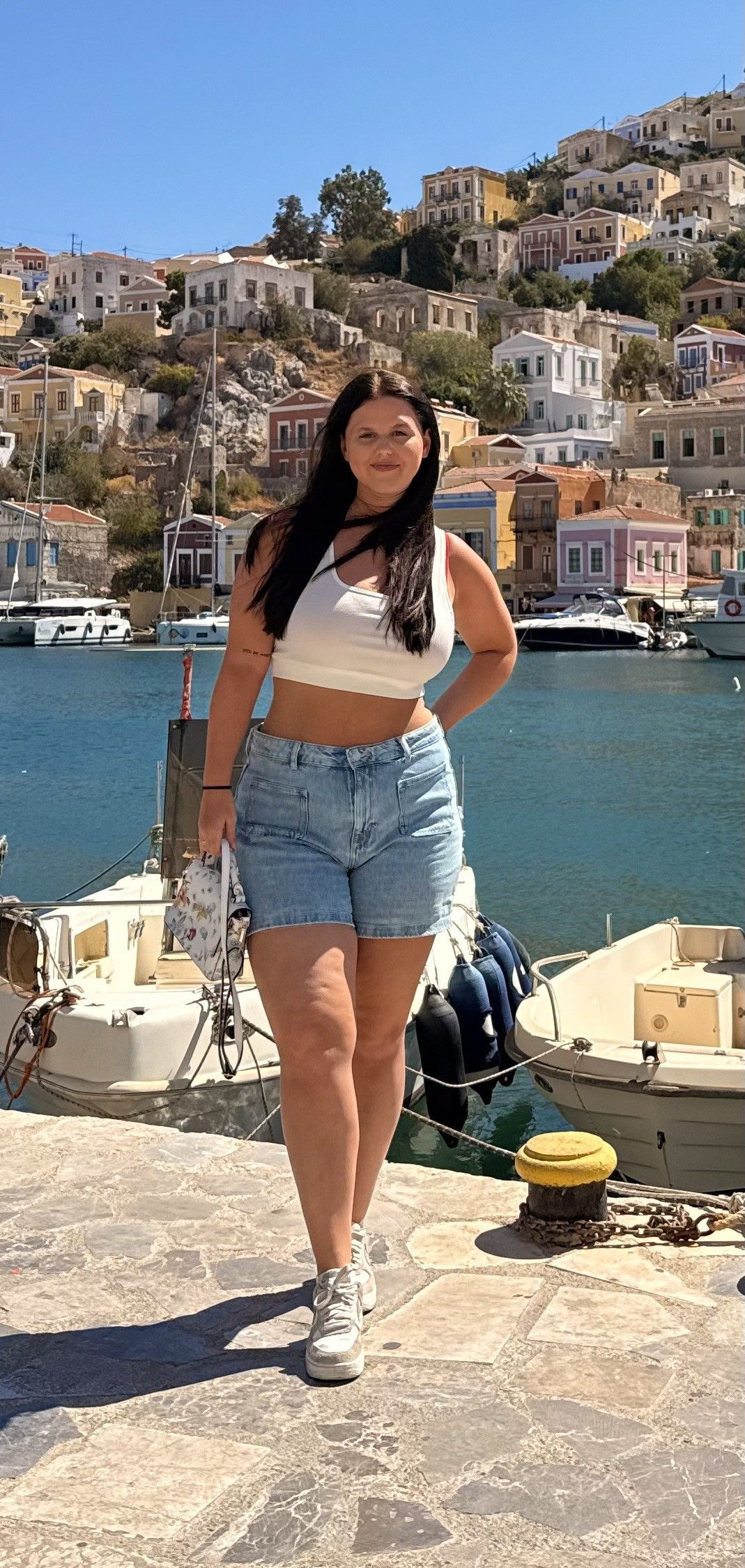 Een vrouw in een spijkerbroekje en crop top poseert bij een haven met gebouwen op een heuvel op de achtergrond.