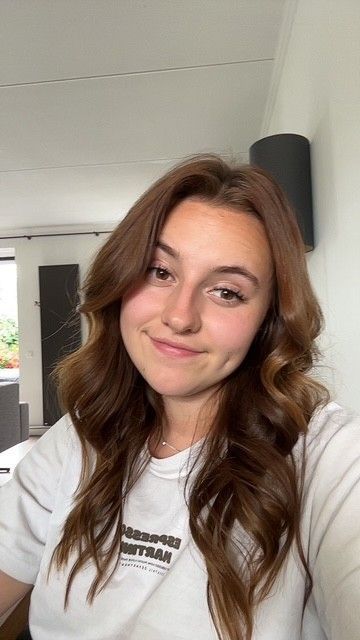 Vrouw met lang, golvend bruin haar, die lichtjes glimlacht. Ze draagt ​​een witte blouse en bevindt zich binnen.