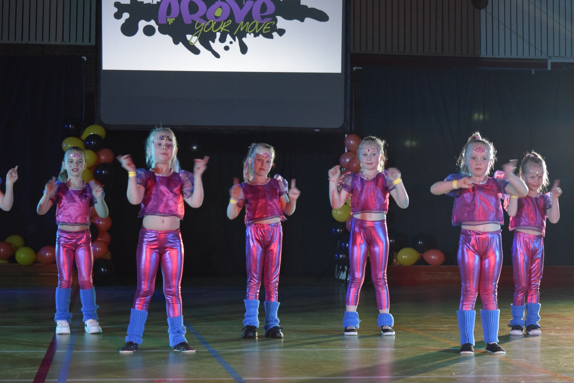 Kinderen in roze outfits dansen op een podium, sommigen steken hun duim omhoog. Op een projectiescherm wordt een logo getoond.