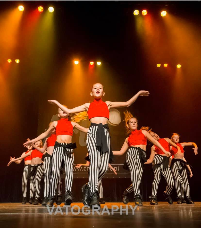 Een groep dansers staat op het podium in rode topjes en zwart-wit gestreepte broeken, met de armen omhoog, onder de podiumverlichting.