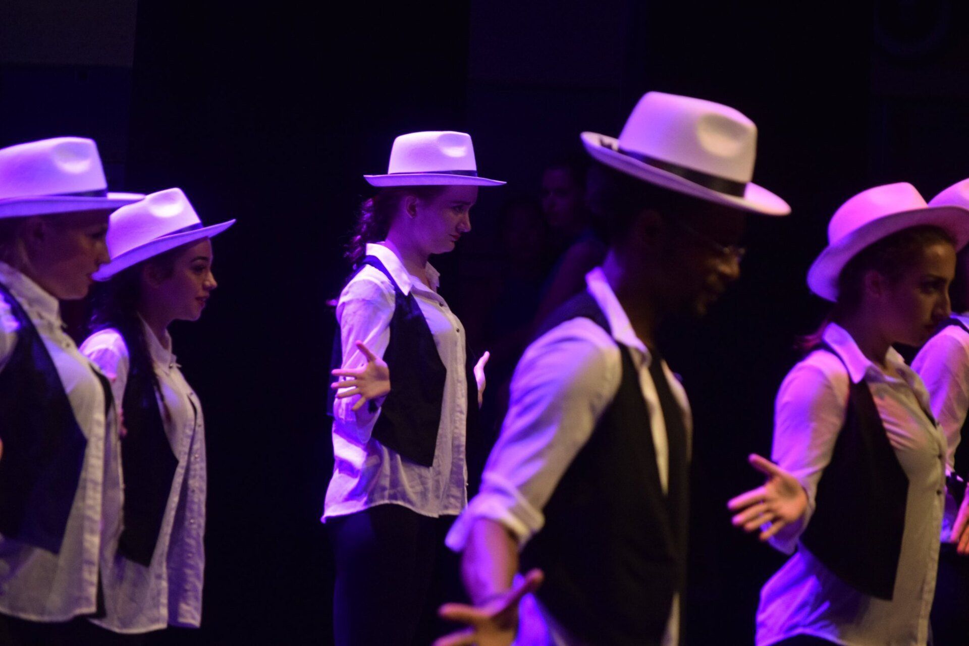 Dansers in witte shirts, vesten en hoeden op het podium. Paarse verlichting.