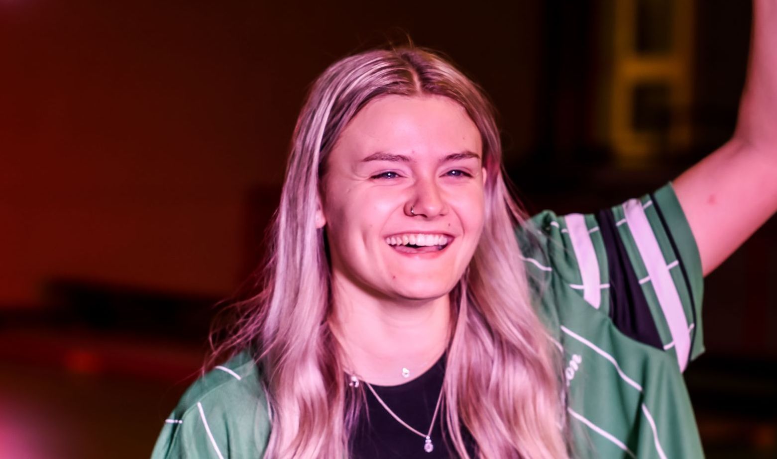 Een glimlachend persoon met lang blond haar, gekleed in een groen-wit gestreept shirt, steekt één arm op in een kamer met warme verlichting.