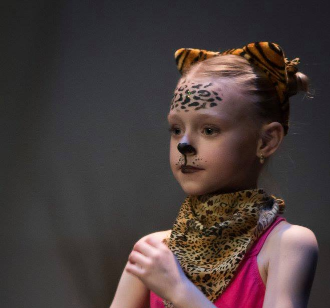 Meisje met cheetah-gezichtsverf, hoofddeksel en nekdoek op het podium, gekleed in een roze turnpakje.