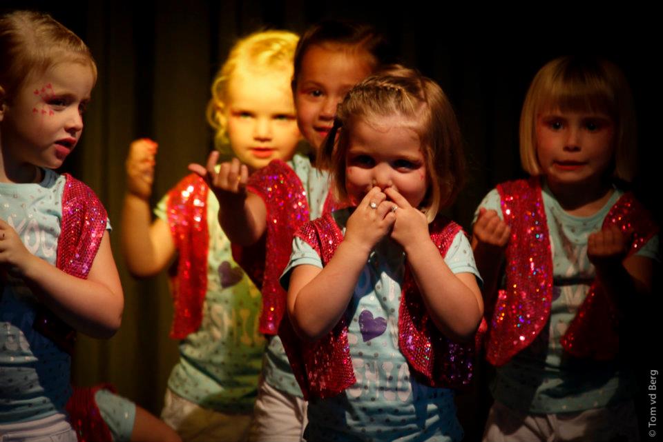 Vijf jonge kinderen in glinsterende vestjes staan ​​op het podium, sommigen met hun handen omhoog en lachend.