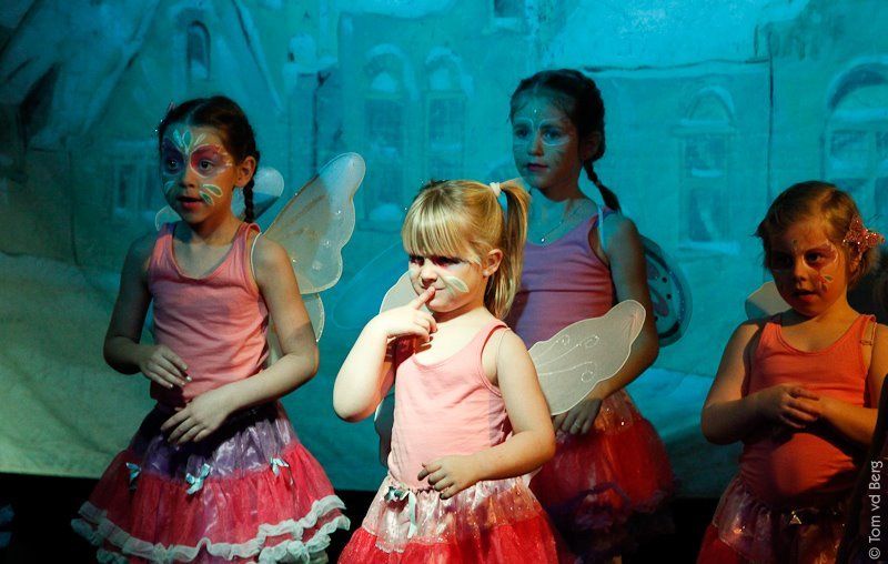 Vier kinderen in feeënkostuums met geschminkte gezichten staan ​​op het podium; een van hen is aan het nadenken.