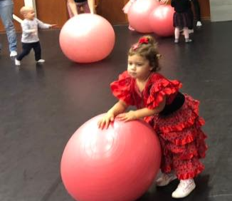 Een meisje in een rode jurk met ruches poseert naast een grote roze bal; op de achtergrond spelen peuters met oefenballen.