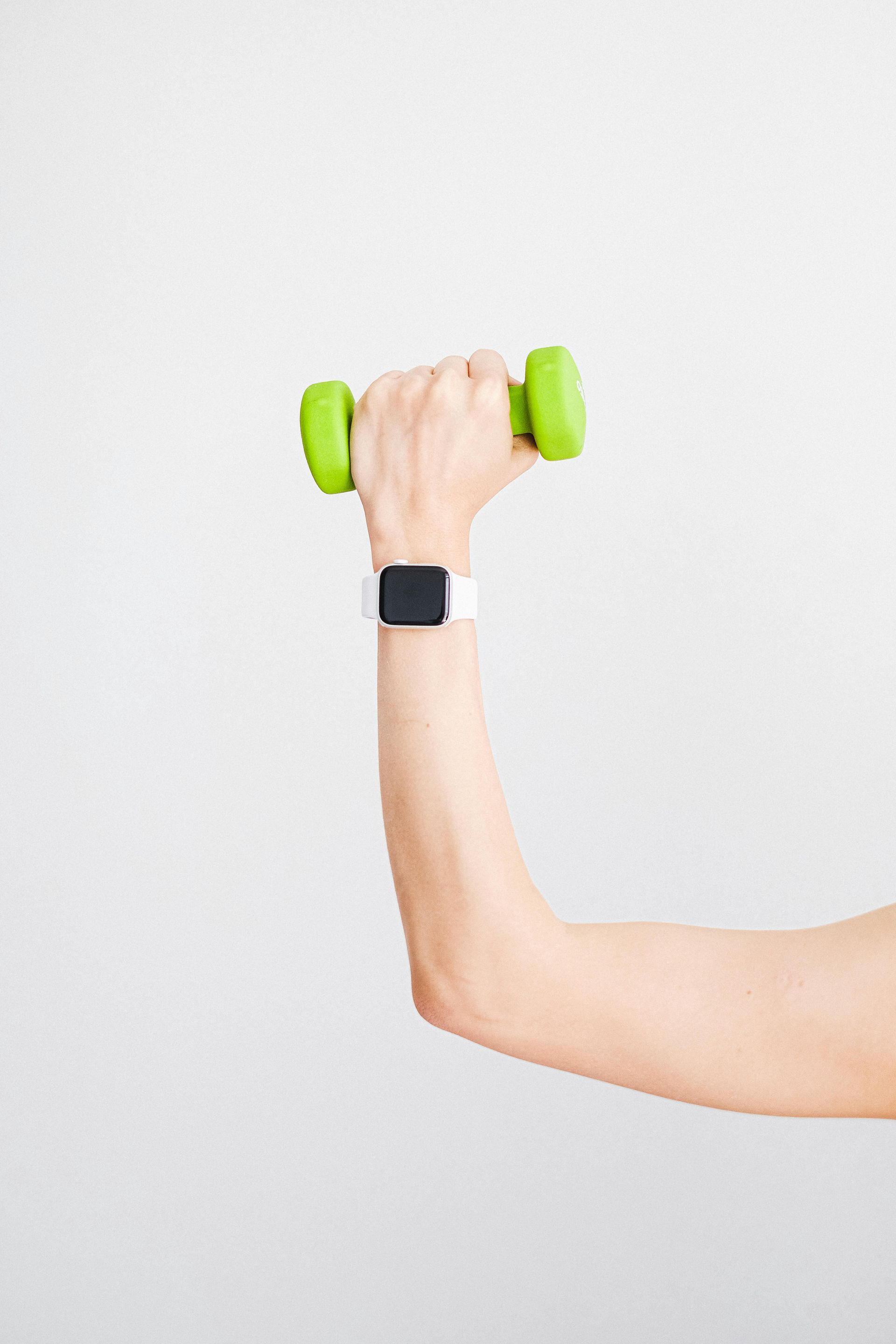 Een arm die een groene halter vasthoudt, draagt ​​een witte smartwatch en staat voor een witte achtergrond.