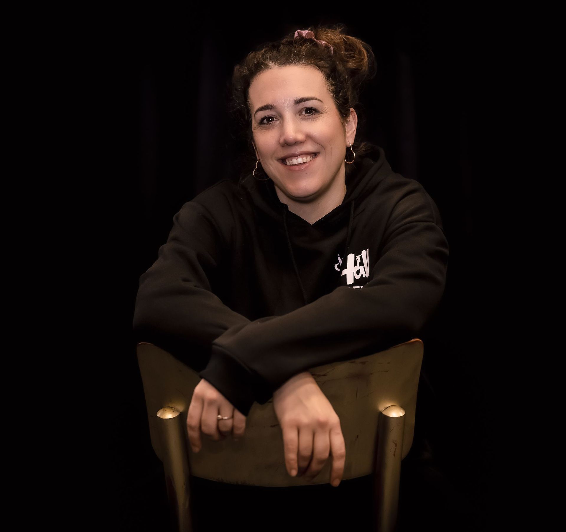 Vrouw in zwarte hoodie glimlacht, zittend met haar armen over elkaar op de rugleuning van een stoel; zwarte achtergrond.