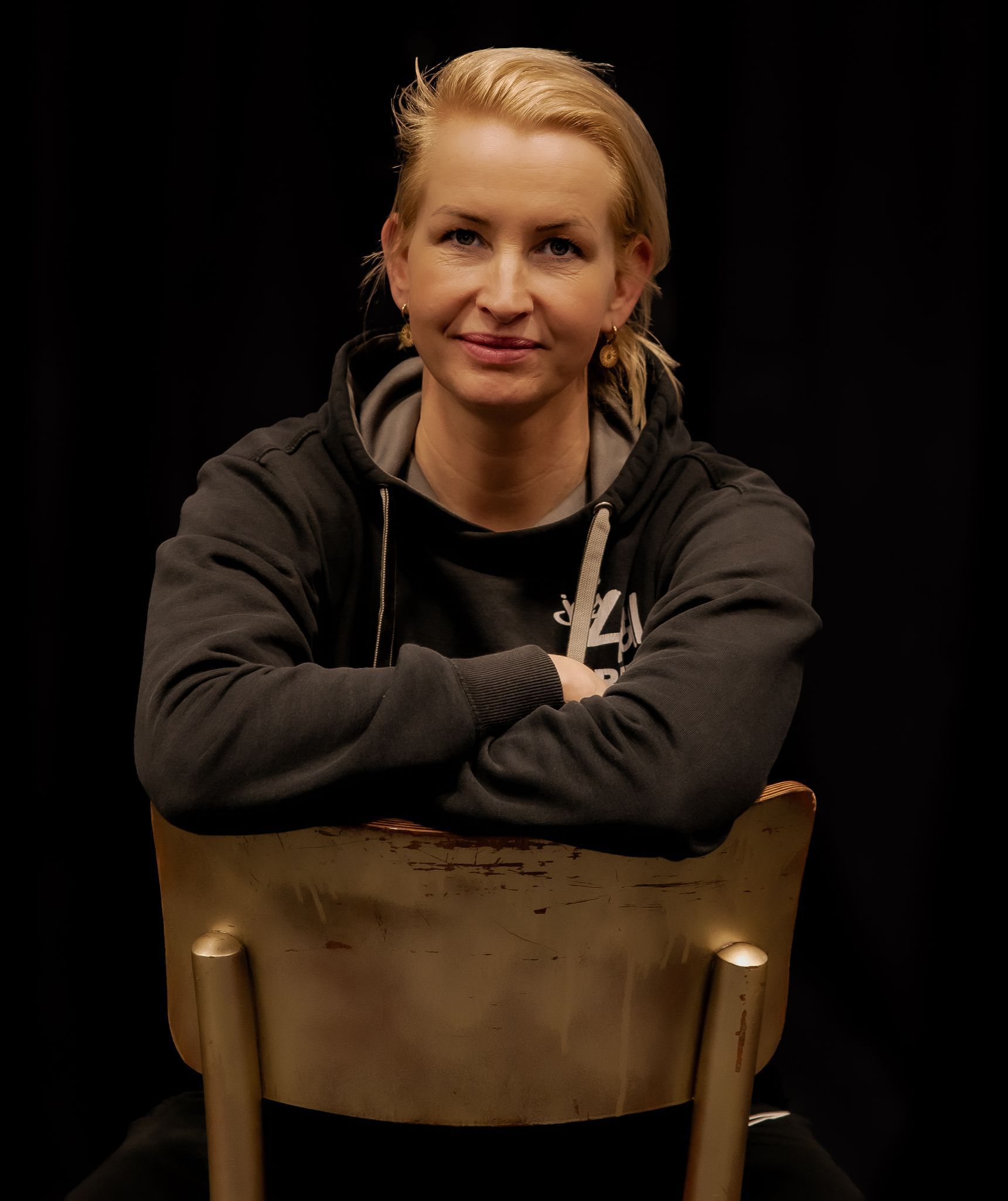 Vrouw in zwarte hoodie zit op een stoel, met de armen over elkaar, en lacht. Donkere achtergrond.
