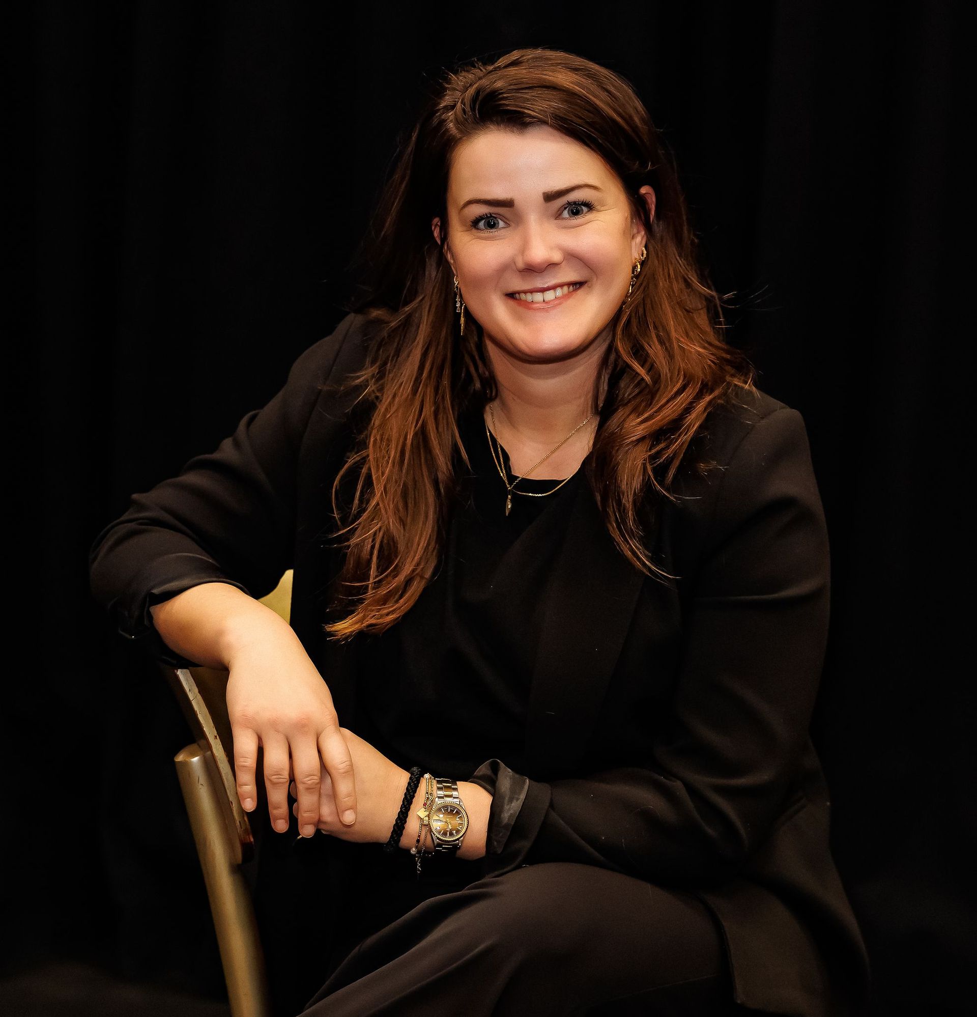 Vrouw met bruin haar glimlacht, zittend in een zwarte blazer en met sieraden; donkere achtergrond.