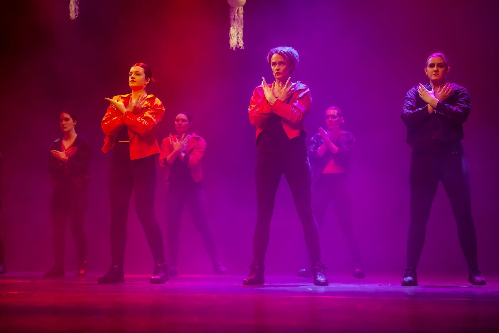 Dansers op een podium in rode jassen en zwarte broeken, met de armen over elkaar. Paarse en rode verlichting.