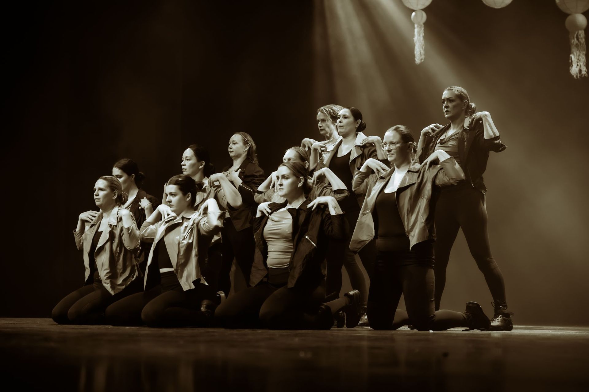 Dansers op het podium, knielend en staand. Sommigen dragen jassen, schijnwerpers boven hen. Sepiatint.