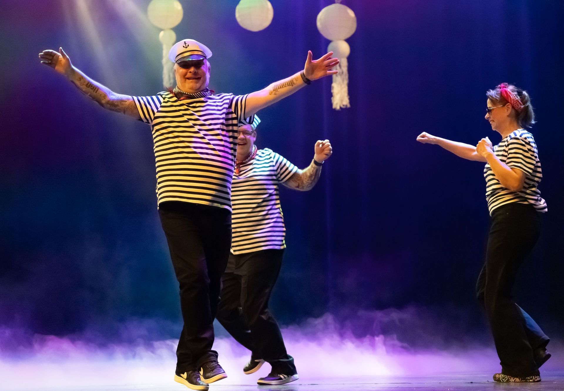 Drie mensen in gestreepte shirts en zwarte broeken dansen op een podium. Rook, ballonnen en schijnwerpers zijn zichtbaar.