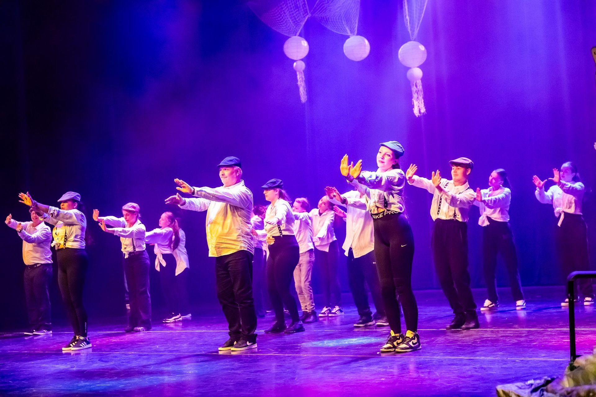 Dansers op een podium met uitgestrekte armen, gekleed in witte shirts, zwarte broeken en blauwe petten, onder paarse podiumverlichting.