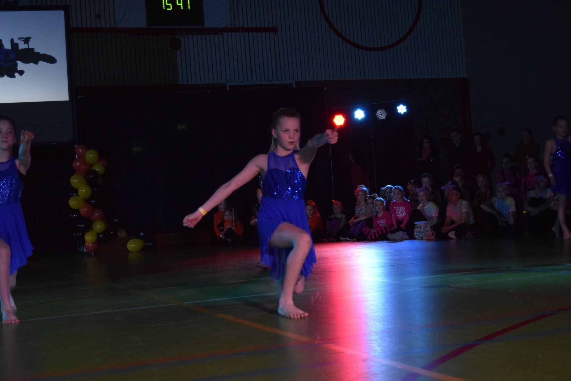 Meisje in een blauwe jurk danst op het podium, met uitgestrekte armen, omringd door kleurrijke lichten en publiek.