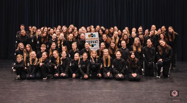 Een grote groep dansers in zwarte kleding staat op het podium en poseert met een bord.
