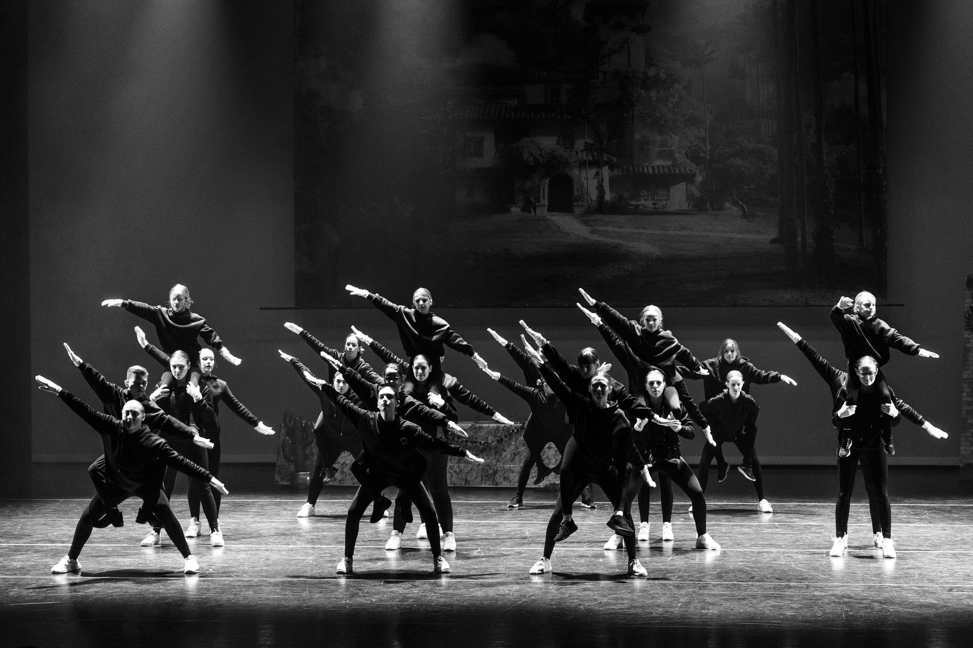 Een groep dansers treedt op het podium op, met de armen omhoog in een synchrone formatie. Zwart-wit.