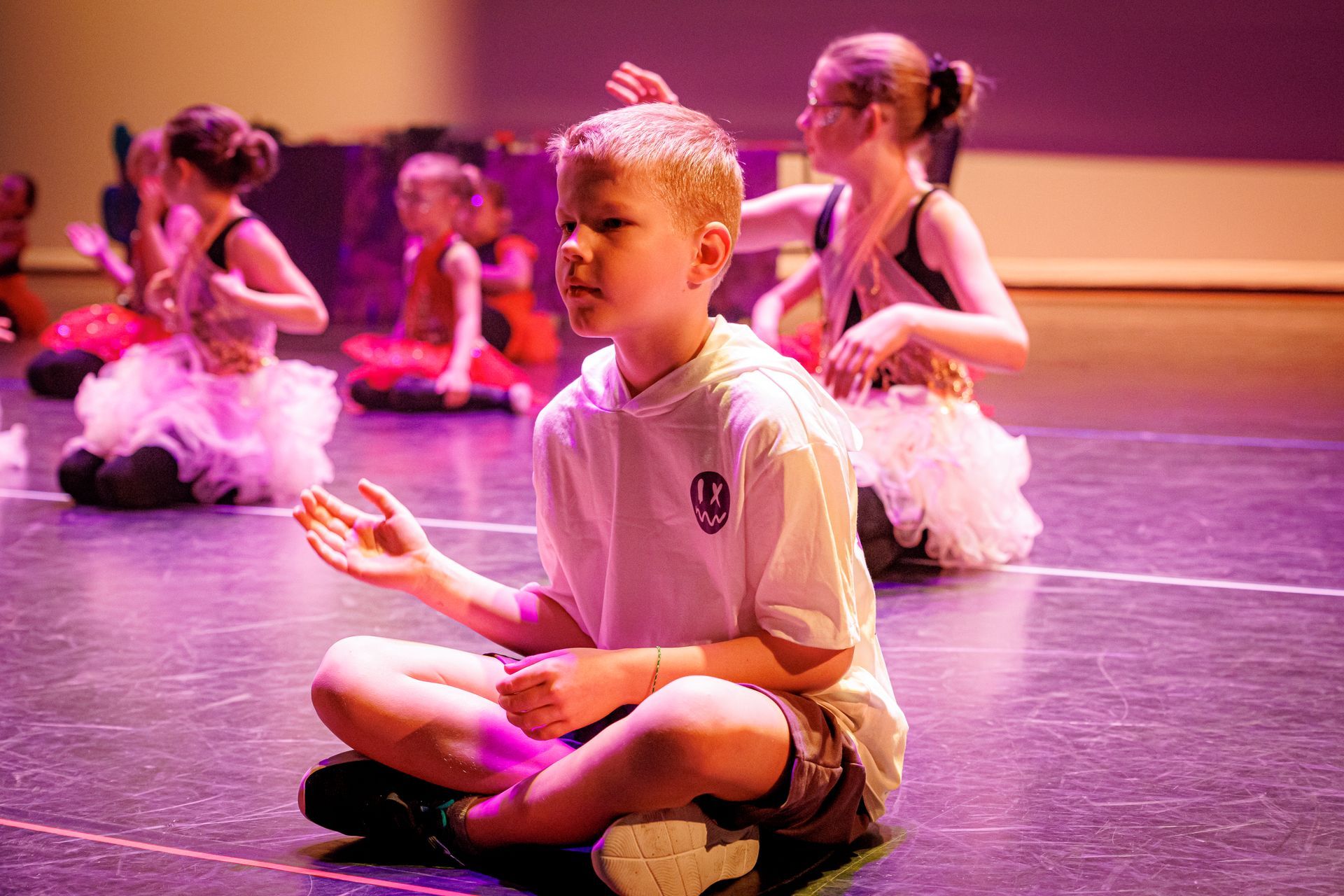 Een jongen mediteert op het podium, met andere dansers op de achtergrond, alles verlicht in paars.