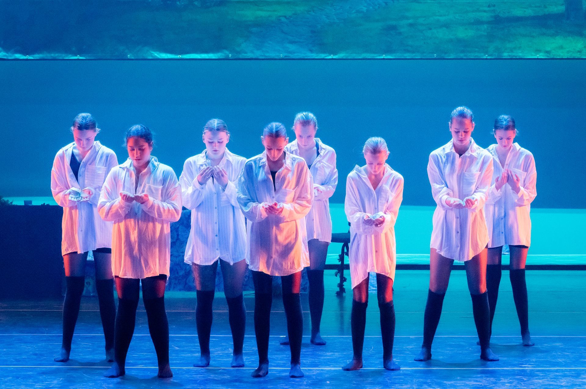 Dansers in witte shirts en zwarte leggings staan ​​hand in hand op het podium, onder blauw licht.