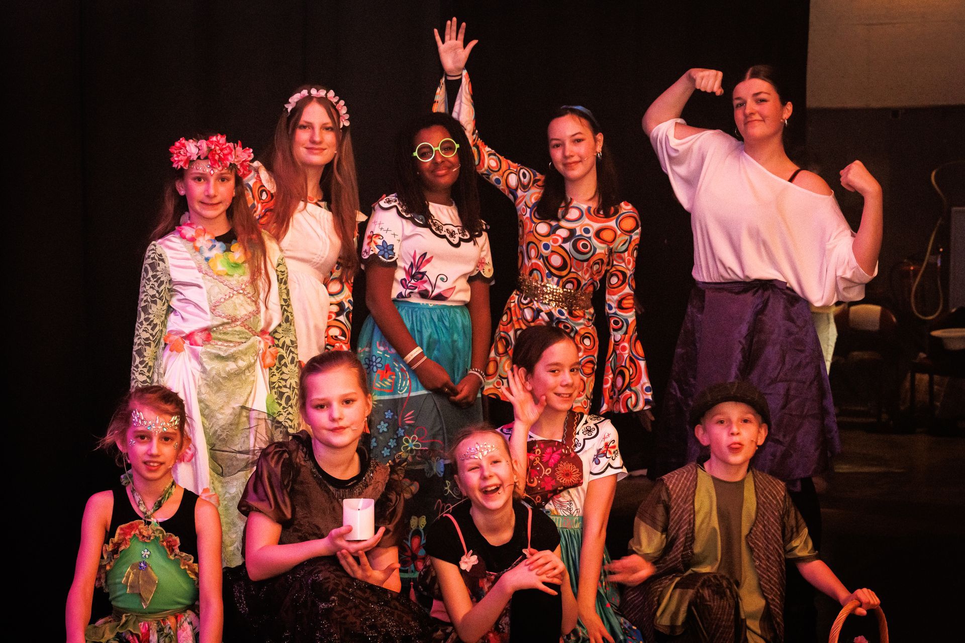 Een groep artiesten in kostuums poseert op het podium met diverse kleurrijke outfits.