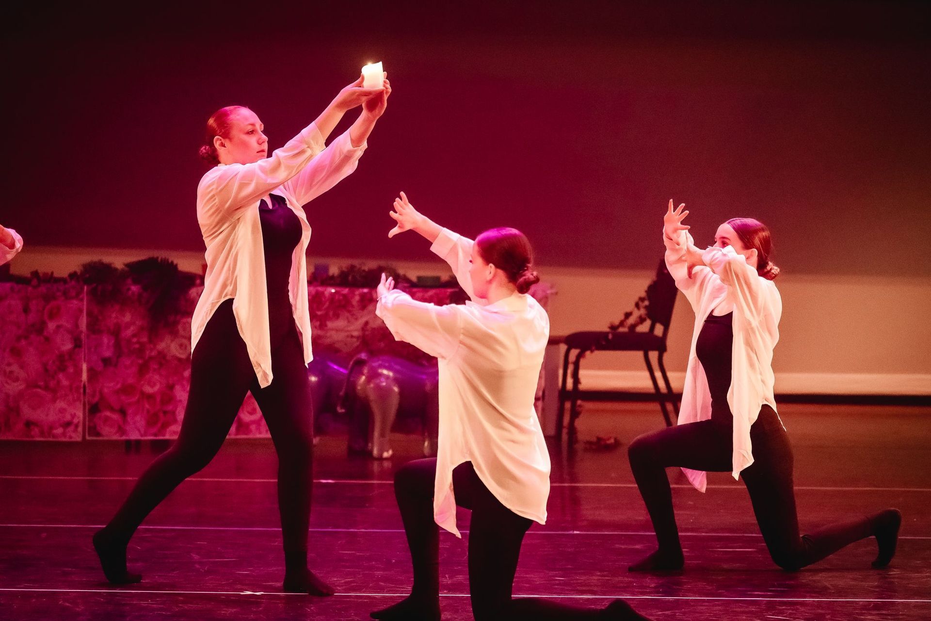 Drie dansers in zwarte leggings en witte shirts houden een brandende kaars op het podium vast en maken er gebaren naar.