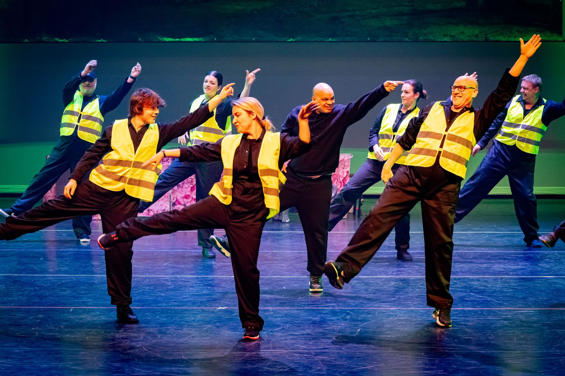 Dansers in zwarte outfits en gele veiligheidsvesten treden op het podium op onder fel licht, waarbij ze hun armen en benen omhoog heffen.